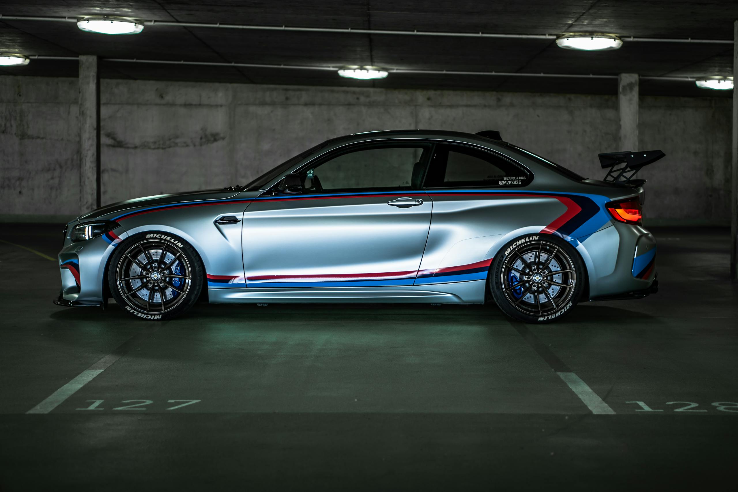 Import Allemagne 1 mandataire automobile limoges achat vente recherche personnalisée import véhicule allemagne limoges gratuit Side view of a BMW M2 sports car with racing stripes, parked in a dimly lit garage.