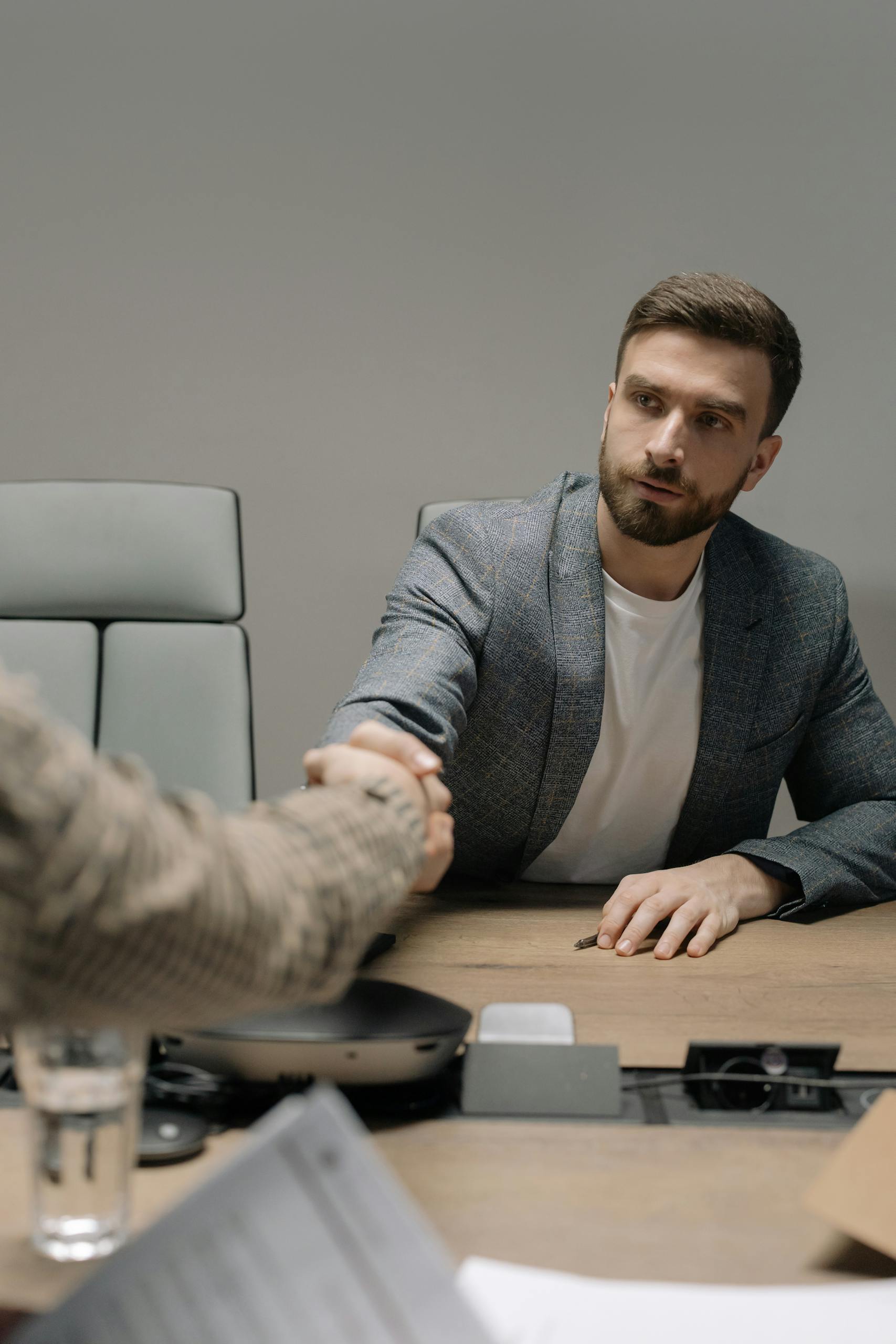 Two businessmen in a modern office shaking hands across a table. Professional setting.